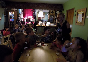 Dzieci siedzą przy stołach i piją sok. Obok dzieci stoi uśmiechnięta kobieta.